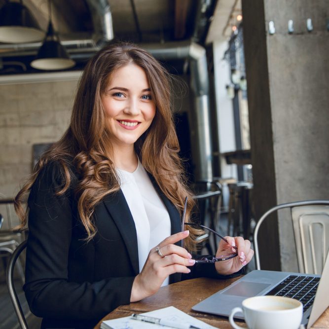 empresaria-muy-sonriente-trabajando-portatil-sentado-cafe Widely smiling businesswoman working on laptop sitting in a cafe. Woman with long hair wears black jacket and white blouse and holds black eyeglasses in hands.